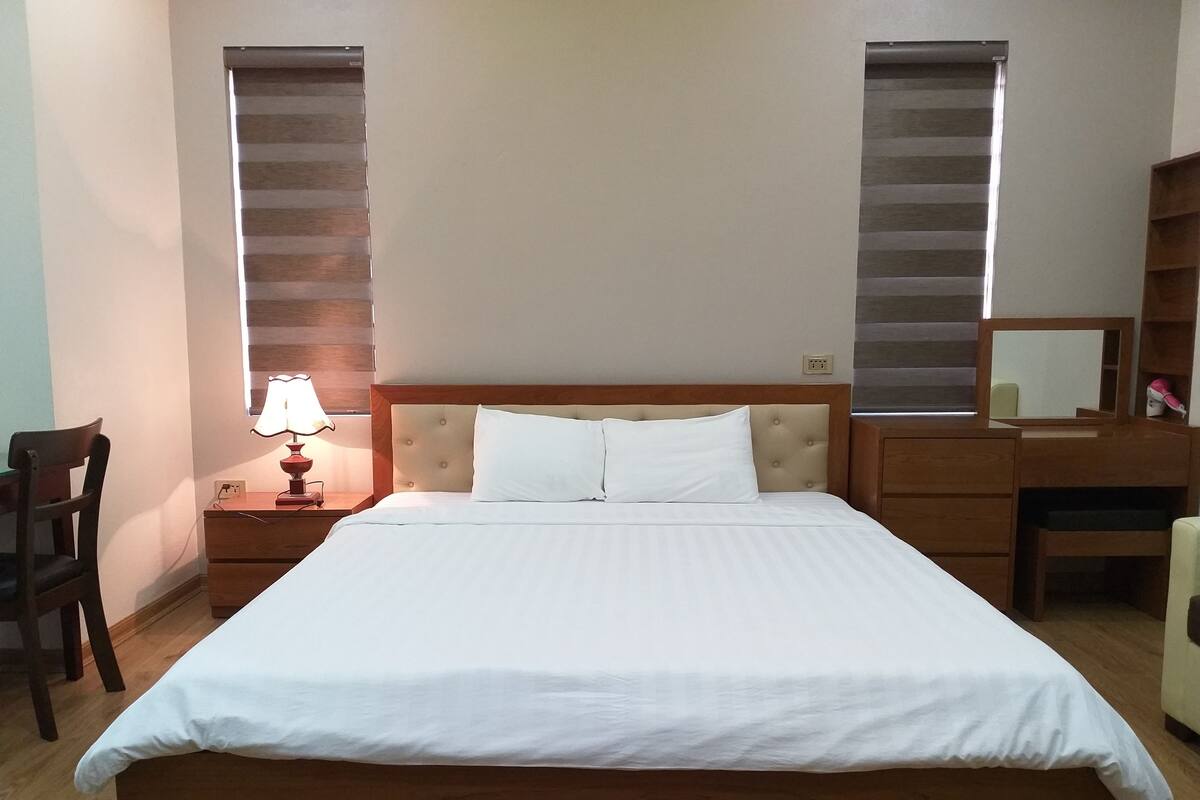 A neatly made bed with a wooden headboard, a bedside table with a lamp, a chair, and a desk with a mirror.