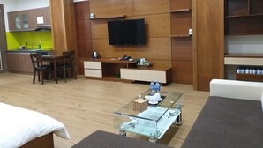 A modern living room with a glass coffee table, a flat-screen TV mounted on the wall, wooden furniture, and a kitchen area with a bright green backsplash.
