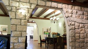 A stone archway leading to a dining area with wooden chairs and a table, a chandelier, and a vase with flowers. - Beit Shalom Historical Boutique Hotel (Metula)