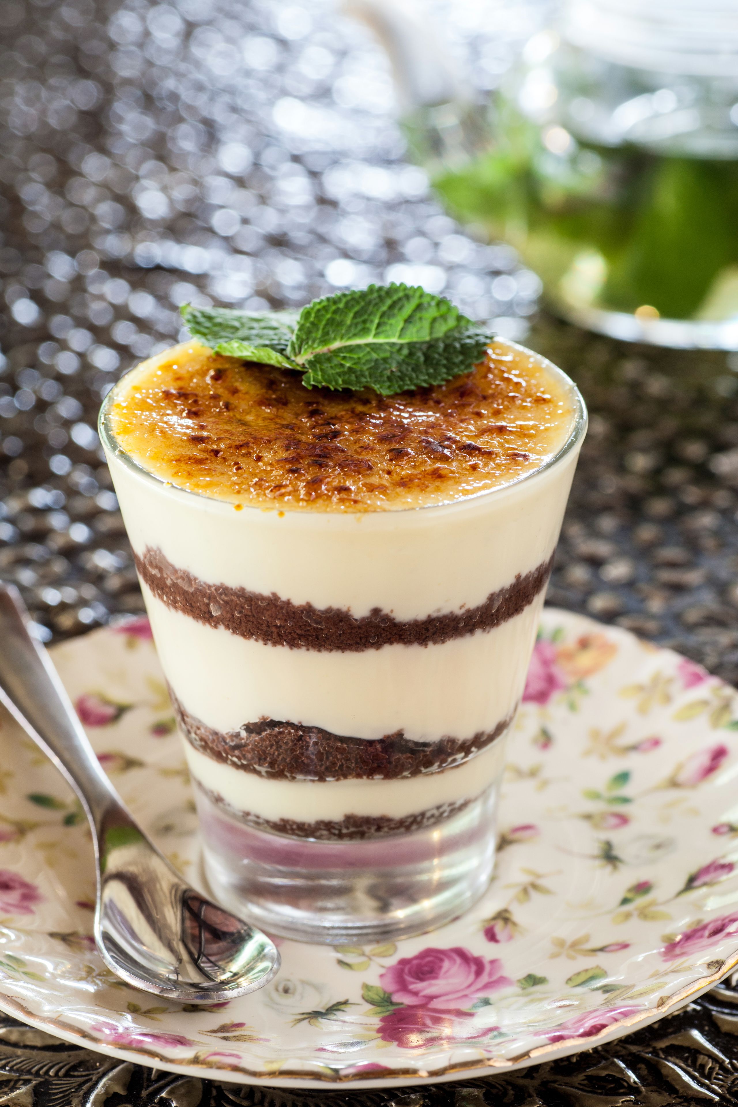 En dessert i flera lager, serverad i ett glas på en tallrik med blommotiv, tillsammans med en sked.