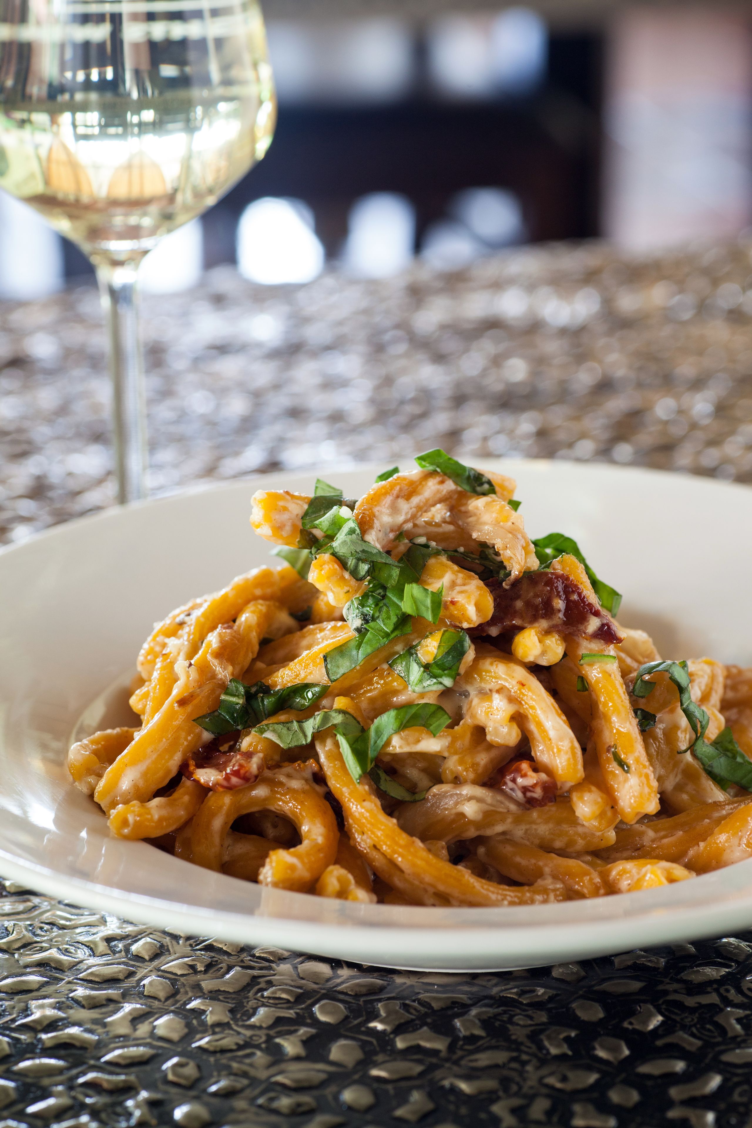 A plate of pasta with chicken and greens, a glass of white wine, and a textured table surface.