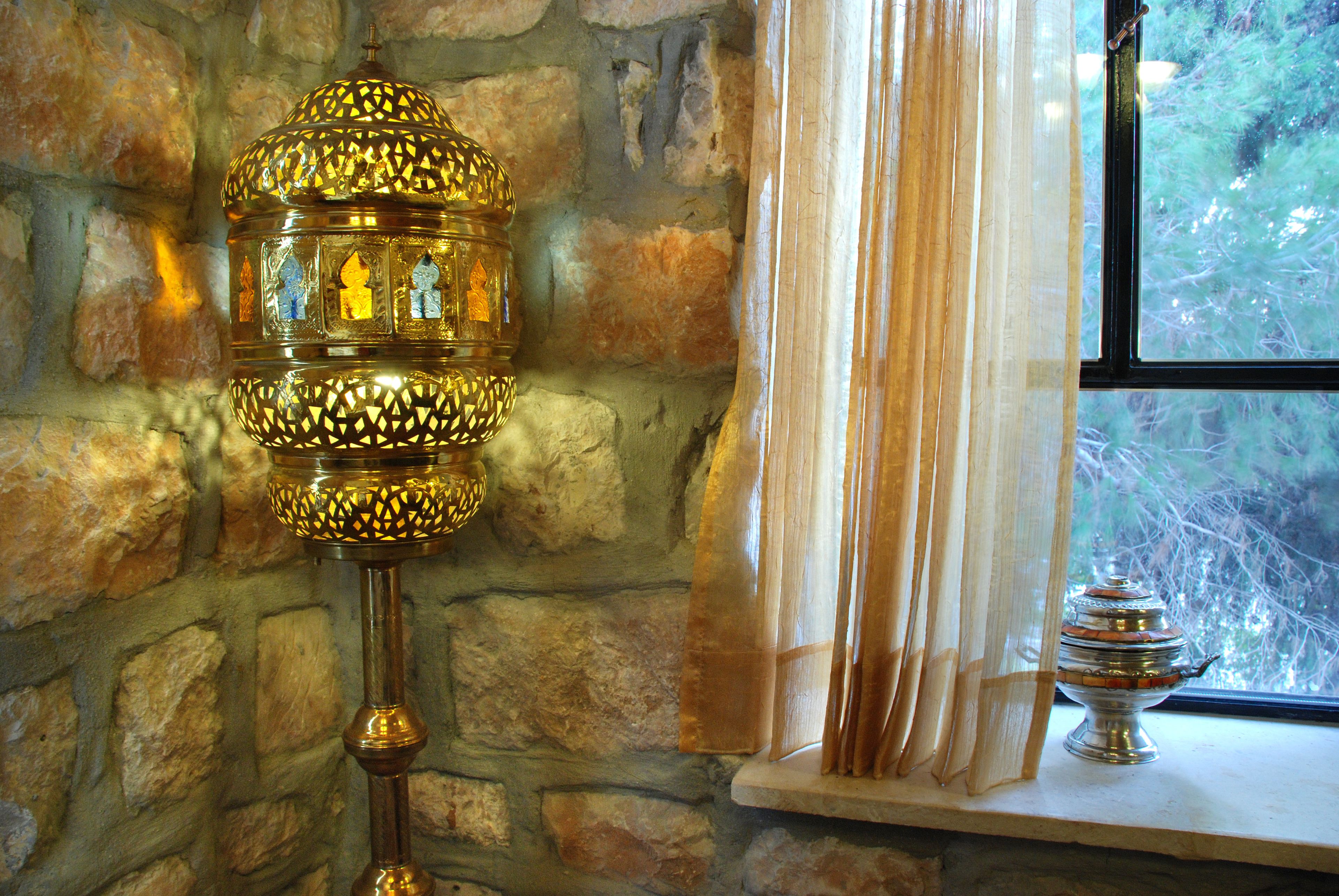 Un mur de pierre, une lampe décorative, une fenêtre donnant sur des arbres et une petite table avec un récipient.