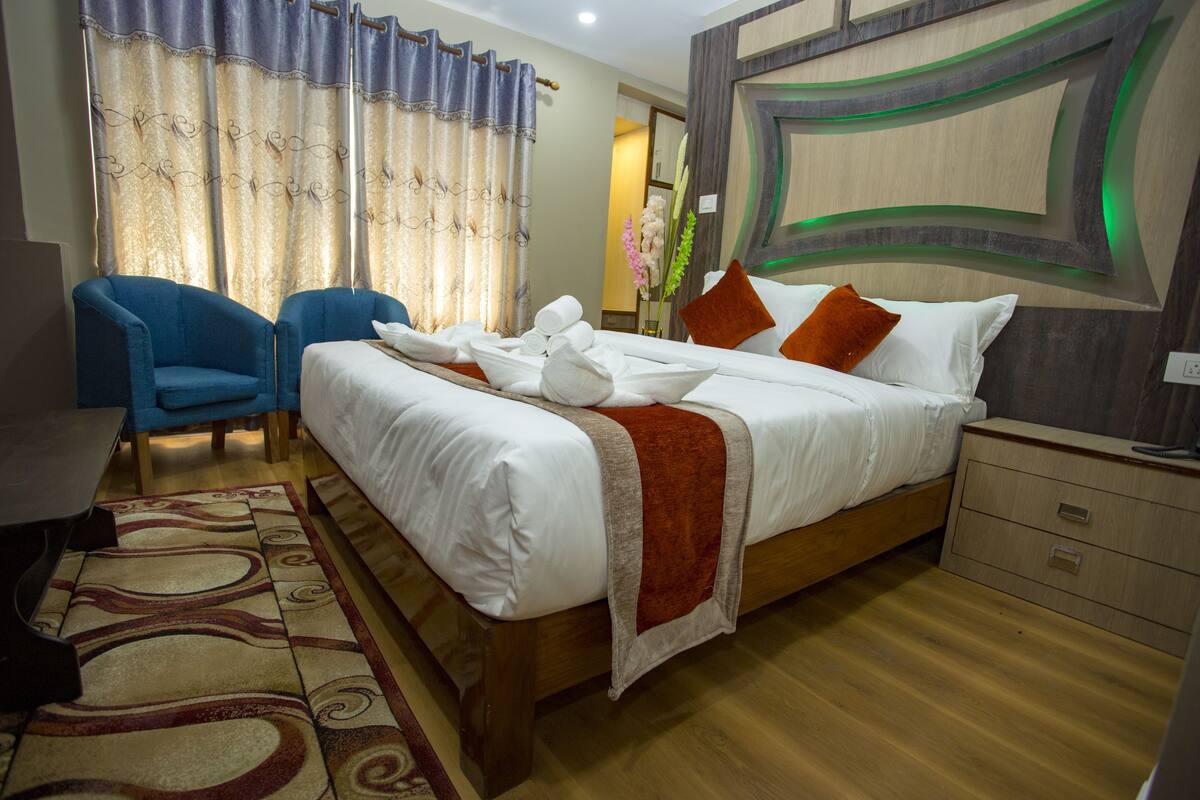 A neatly made bed with white linens and a red throw blanket, a blue armchair, a wooden nightstand with a lamp, and a decorative wall with a geometric pattern.