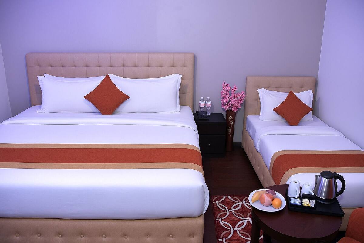 A hotel room with two beds, each with white bedding and a brown headboard. There is a small table with a tray of fruit and a kettle.