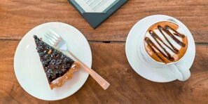 A slice of blueberry cheesecake on a white plate with a fork, and a cup of coffee with chocolate drizzle on a saucer. - Hotel Marinha (Kathmandu)