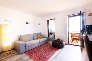 A living room with a sofa, a red patterned rug, a bag, and a door leading to a balcony.
