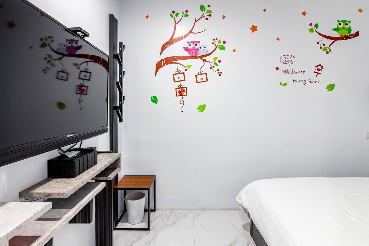 A bedroom with a TV mounted on the wall, a bed, a small wooden table, and a trash bin.
