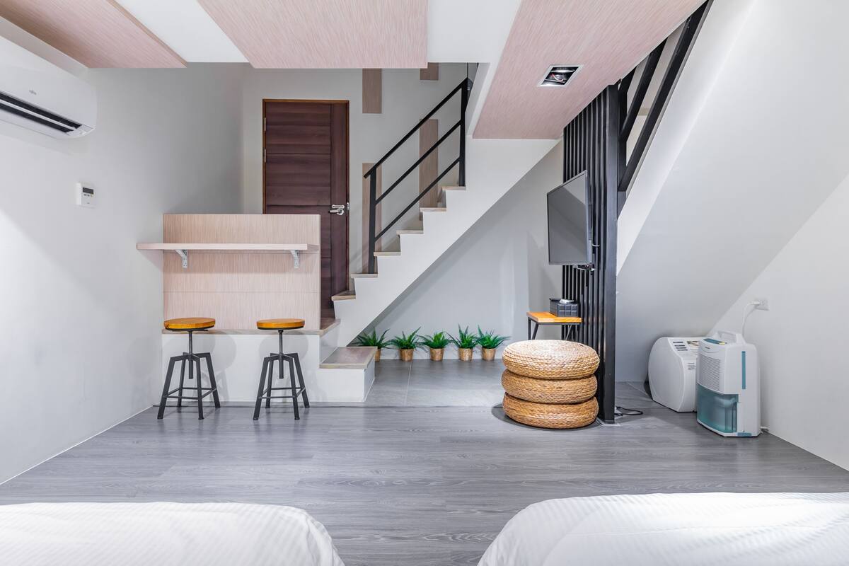 A modern interior with a staircase, a small seating area with stools, a round wicker ottoman, and a white air purifier.