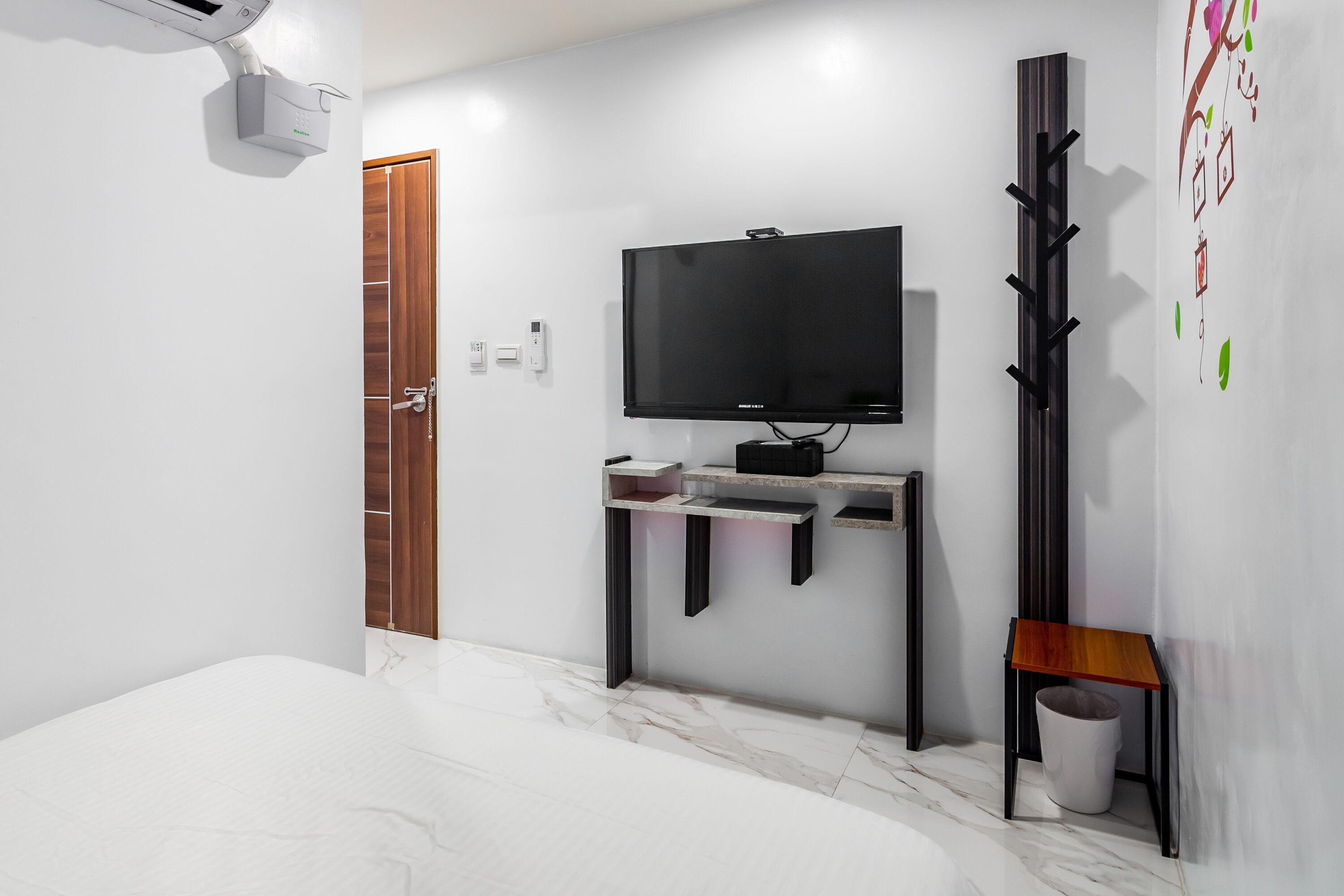 Een moderne slaapkamer met een bed, een aan de muur bevestigde televisie, een kapstok en een kleine prullenbak.