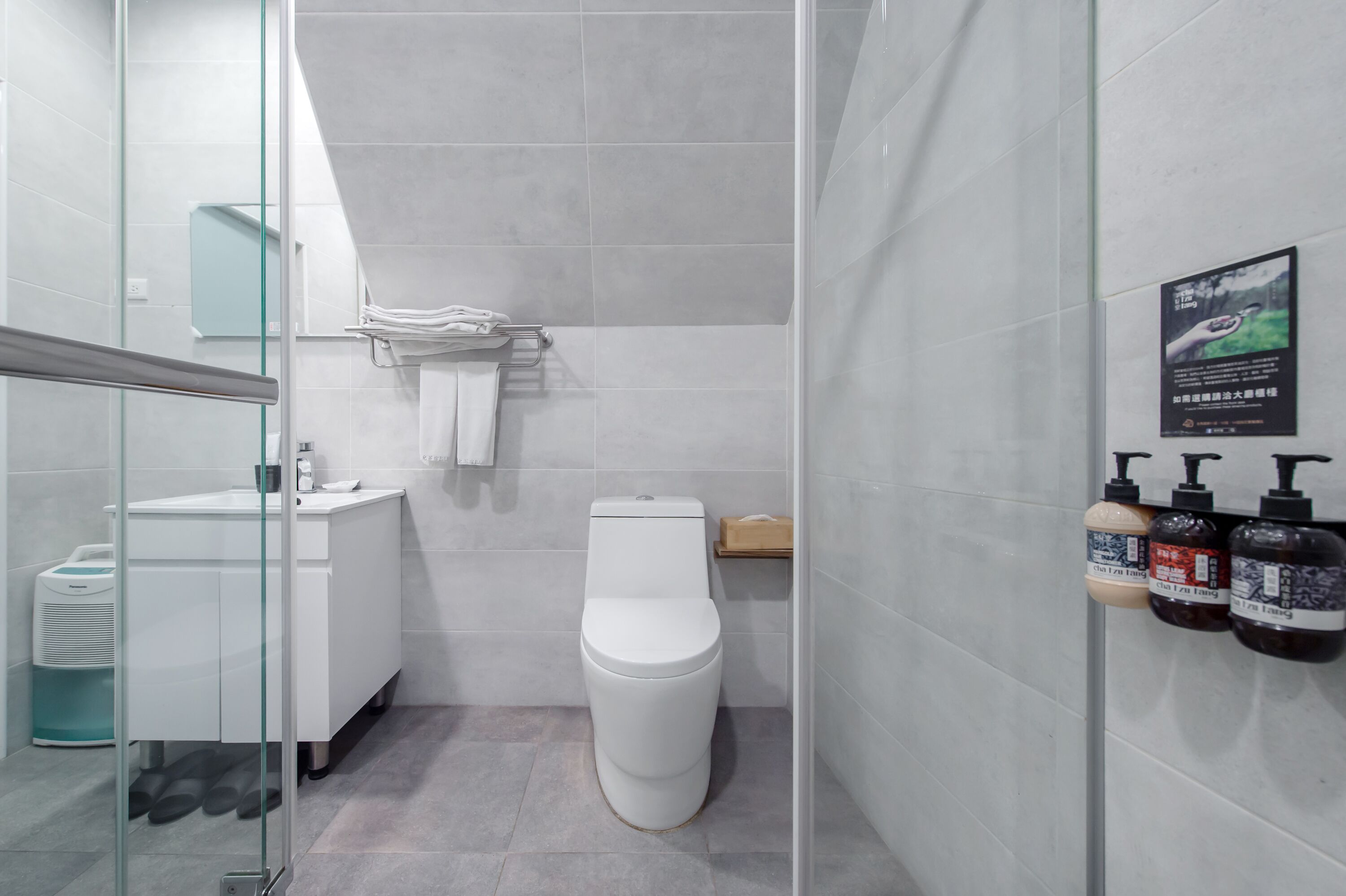 Un baño moderno con cabina de ducha de vidrio, inodoro blanco, lavamanos con espejo y estante con artículos de aseo.