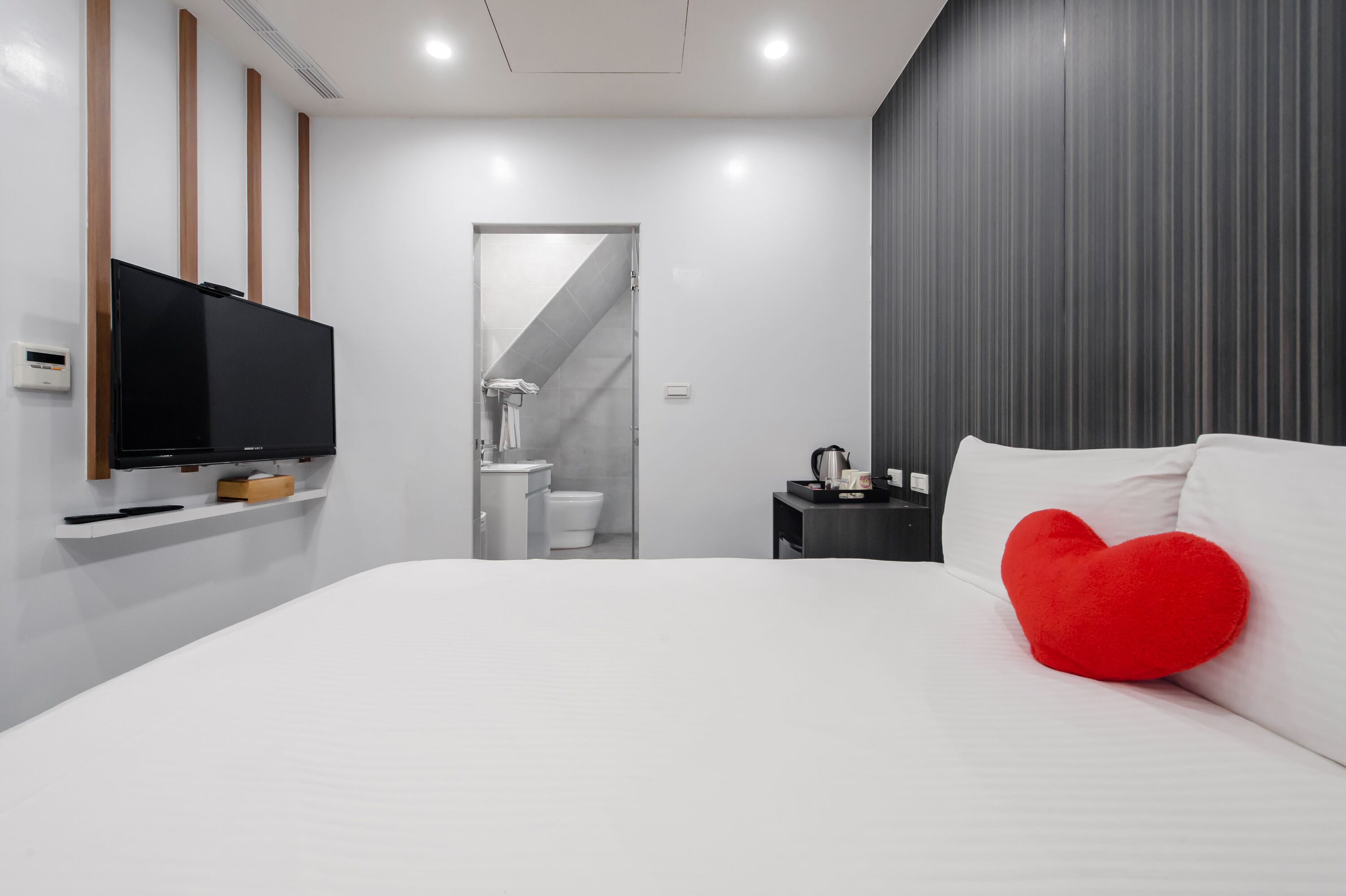 Un dormitorio moderno con una cama grande, un televisor montado en la pared, un cojín rojo con forma de corazón y vistas al baño.