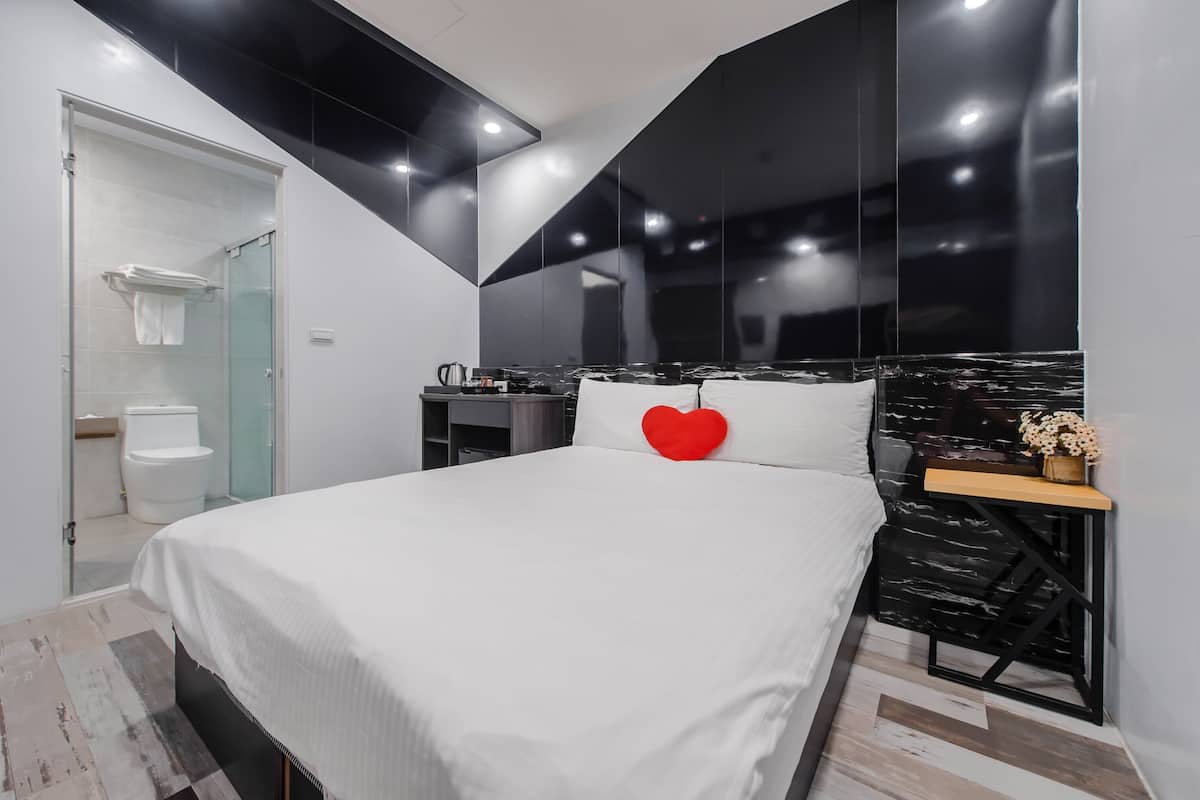 A modern bedroom with a bed, a red heart-shaped pillow, a nightstand with a vase, and a view of the bathroom.
