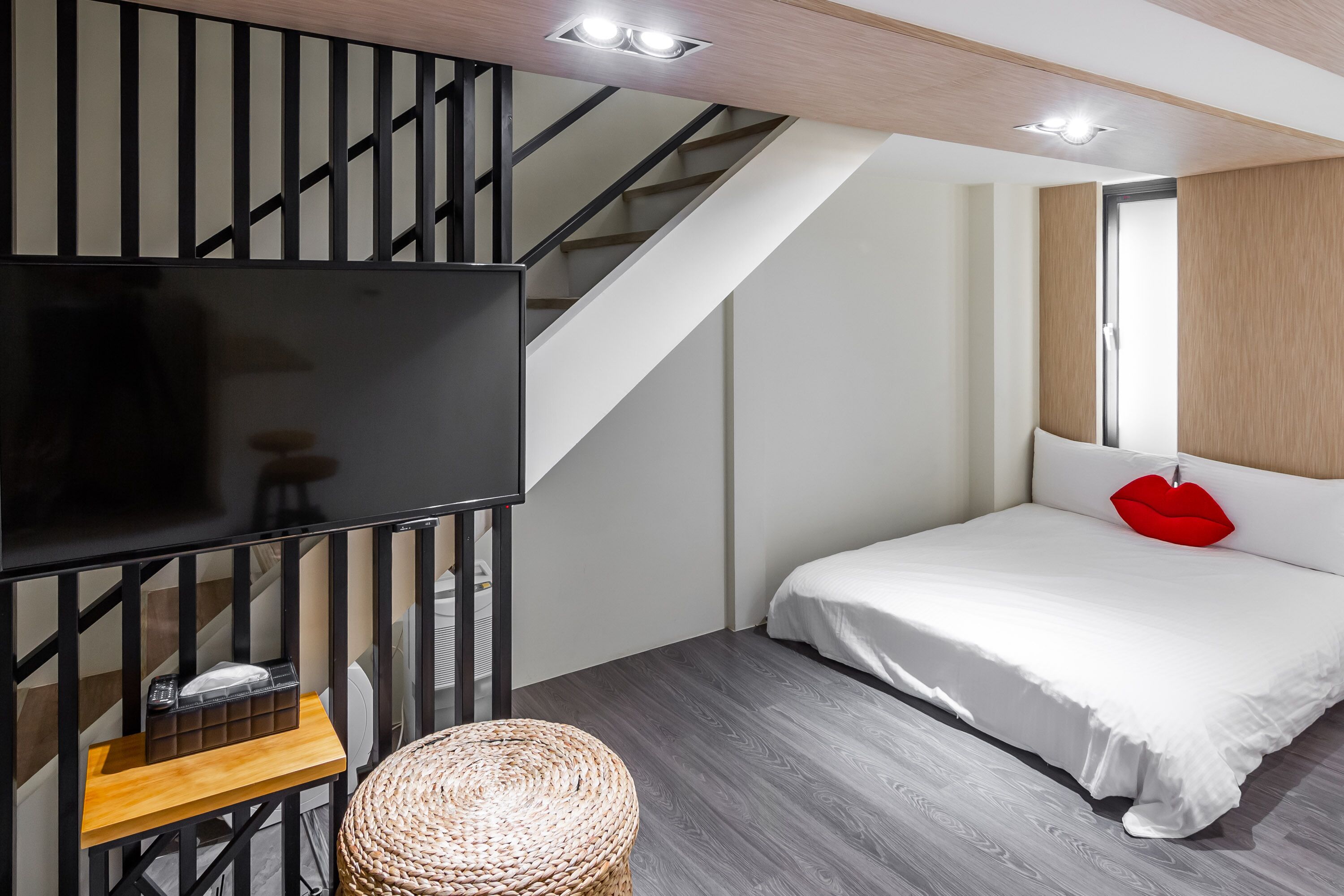 A modern bedroom with a large TV, a bed with white bedding and a red pillow, a woven stool, and a staircase with black railings.