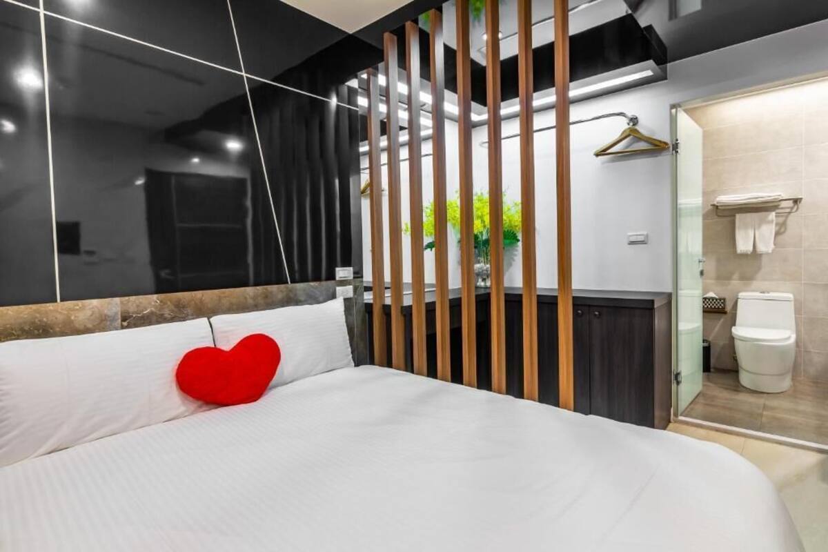 A modern bedroom with a large bed, a red heart-shaped pillow, a wooden partition, and a view of the bathroom.