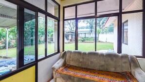 Quarto com sofá e janelas amplas que oferecem vista para um jardim e um edifício de tijolos.
