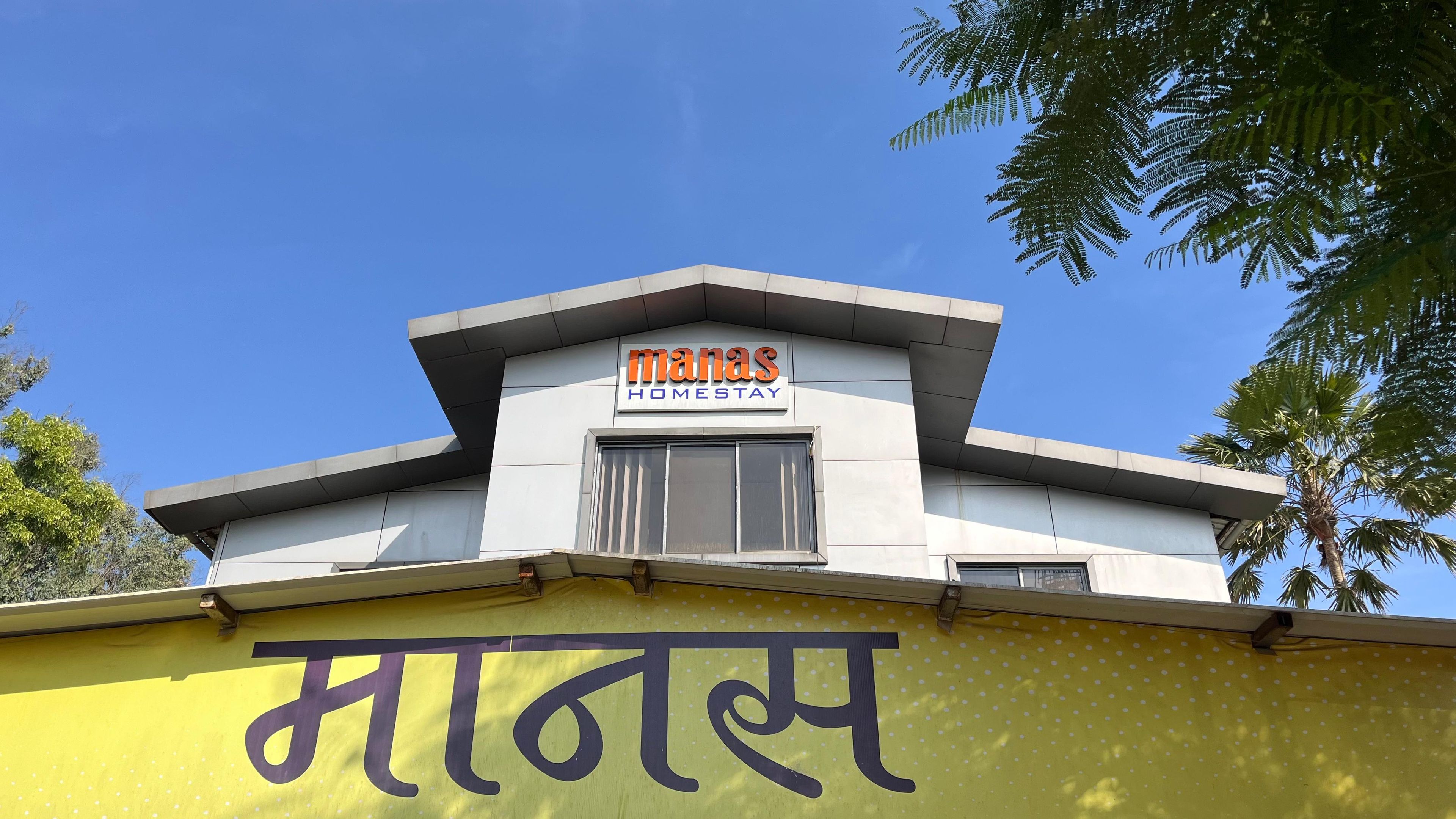 A building with a sign reading 'Manas Homestay' and a large yellow wall with the word 'MANAS' in Hindi.