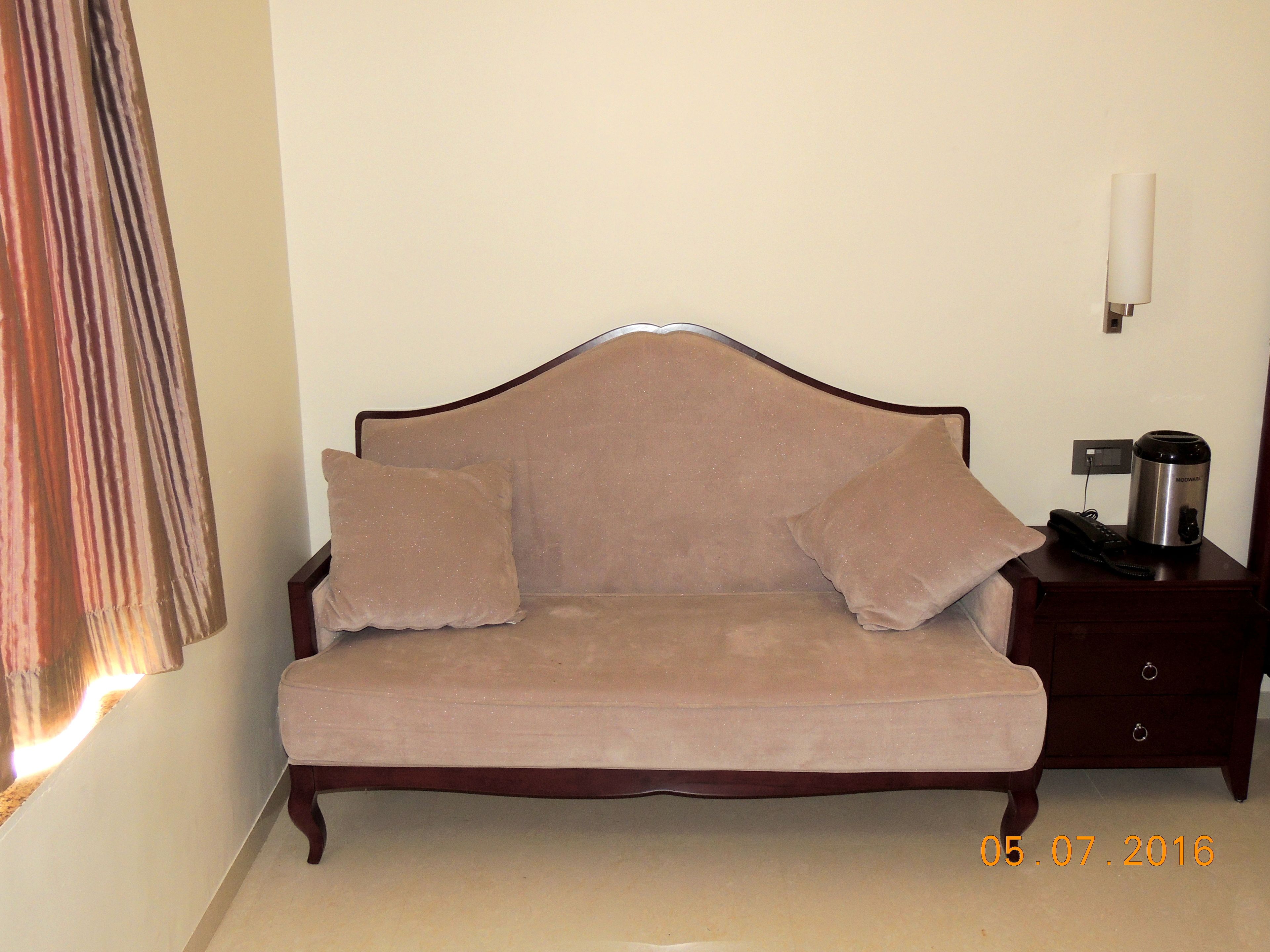 Un sofá beige con dos almohadas, una mesita de noche con cafetera y una lámpara de pared.