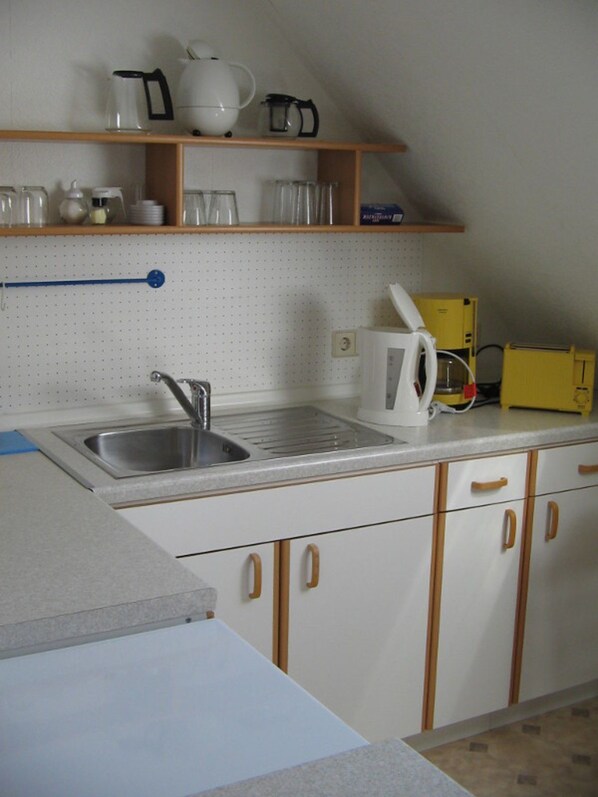 A kitchen with a sink, cabinets, a countertop, and a shelf with kitchenware.