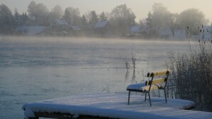 Um banco coberto de neve, um lago congelado e casas ao fundo.