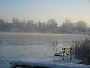 A bench with snow on it, a frozen lake, and houses in the background.