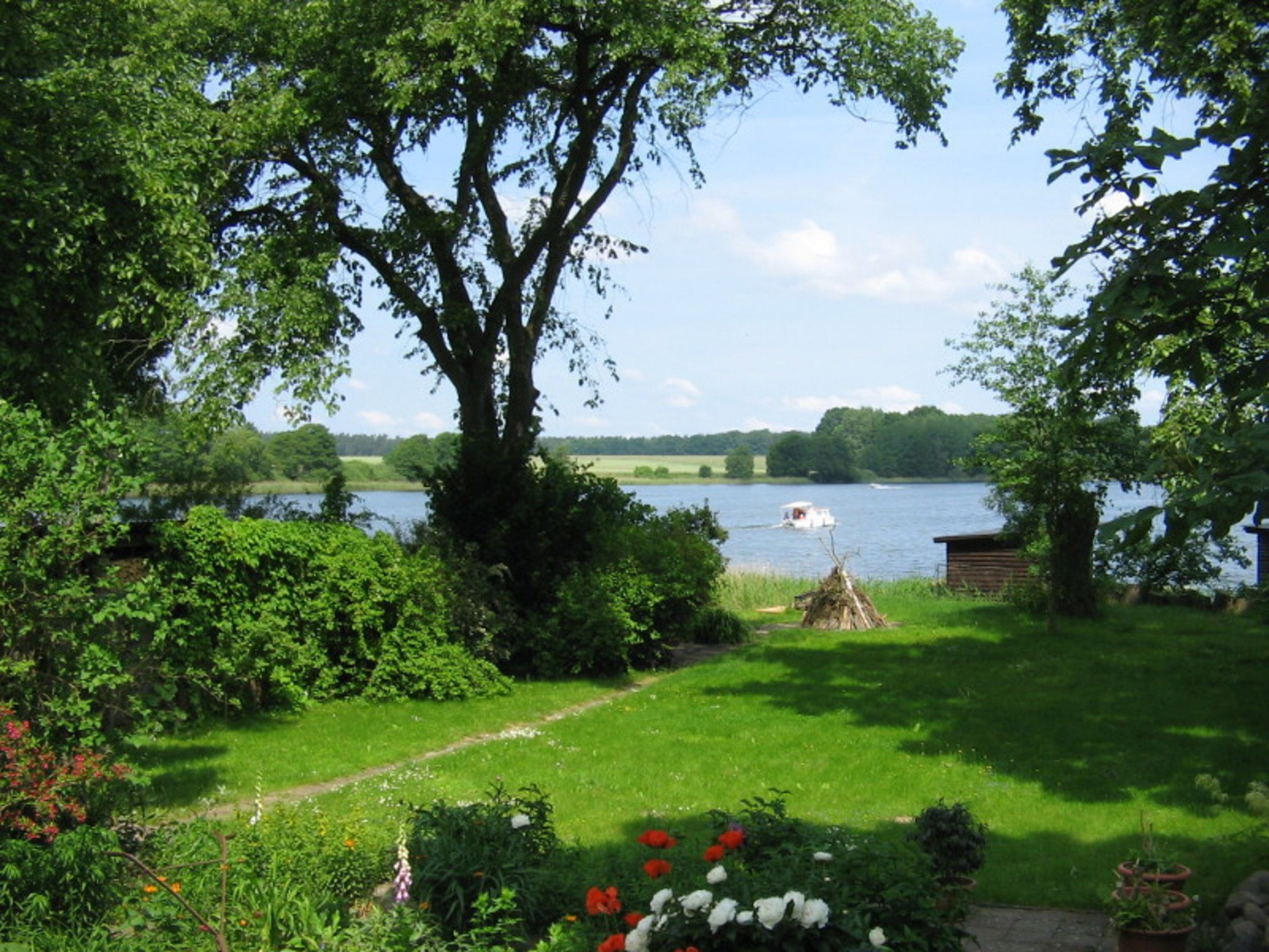 Een tuin met een grote boom, een tuinhuisje en een pad dat naar een meer met een boot leidt.