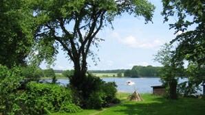 Um jardim com uma grande árvore, um pequeno galinheiro e um caminho que leva a um lago com um barco.