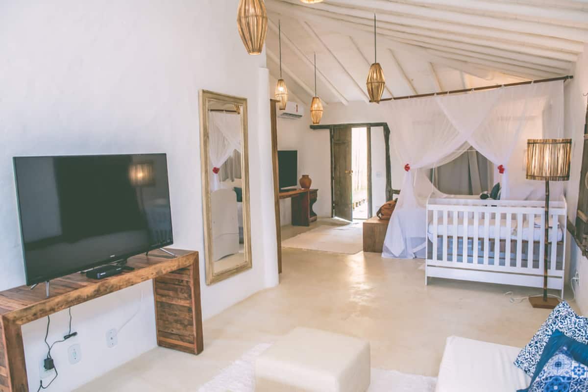 Una habitación con televisión sobre un mueble de madera, una cuna blanca, un espejo y una cama con dosel.