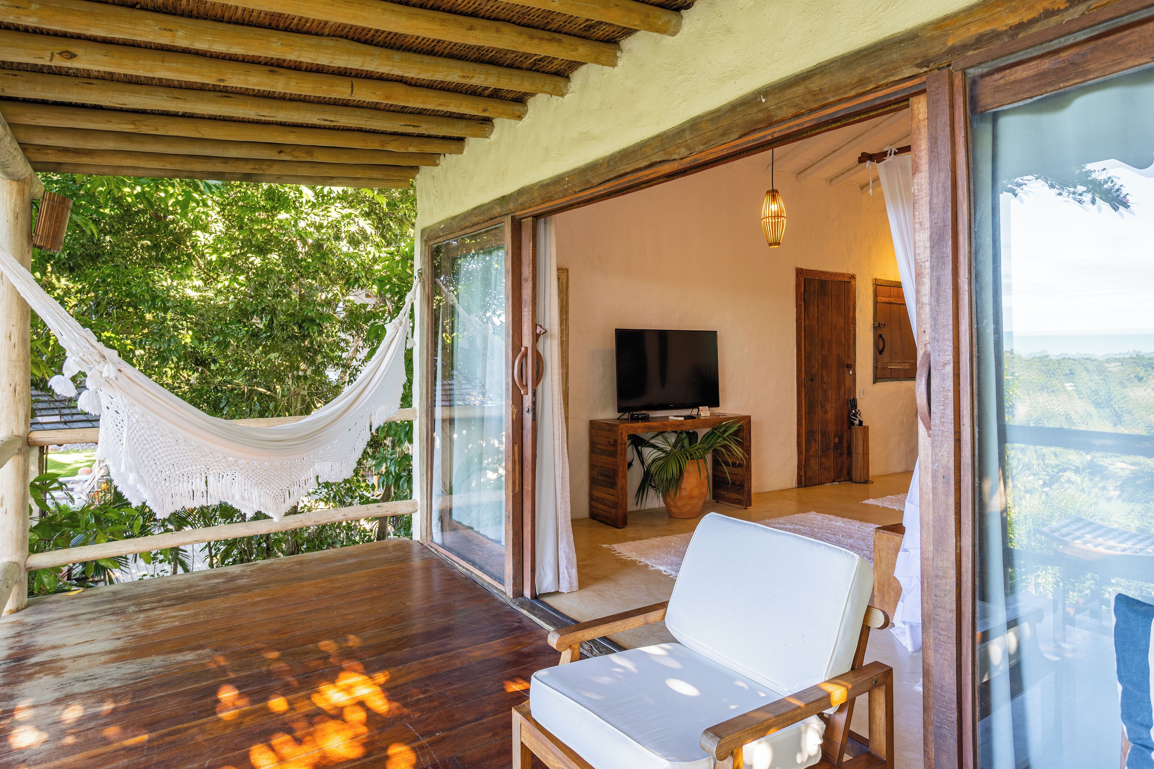Una terraza de madera con una hamaca, un televisor en una habitación con puerta corredera de vidrio y vista a la vegetación.