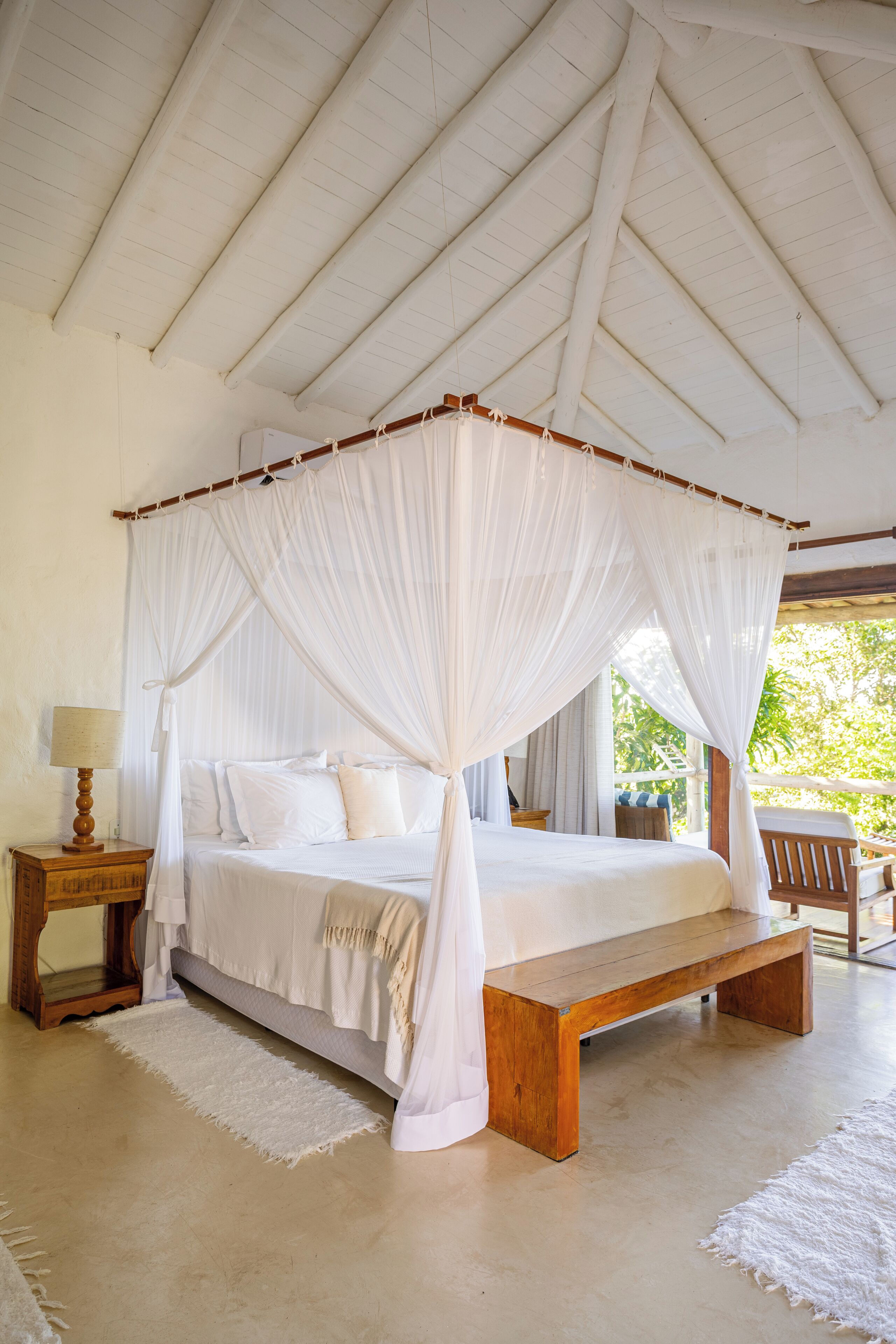 A four-poster canopy bed with white linens, a wooden bench, and a bedside table with a lamp.