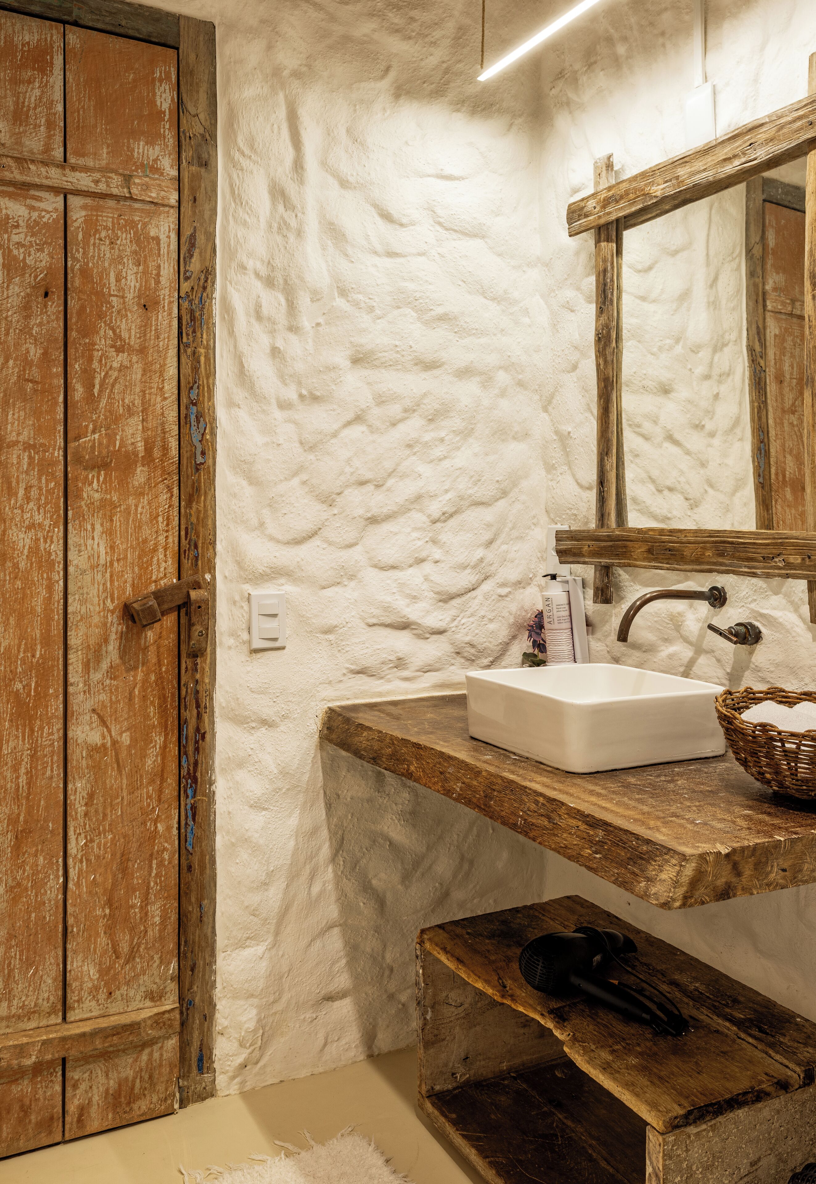 A bathroom with a rustic wooden door, a textured white wall, a wooden countertop with a sink, a mirror, and a basket.