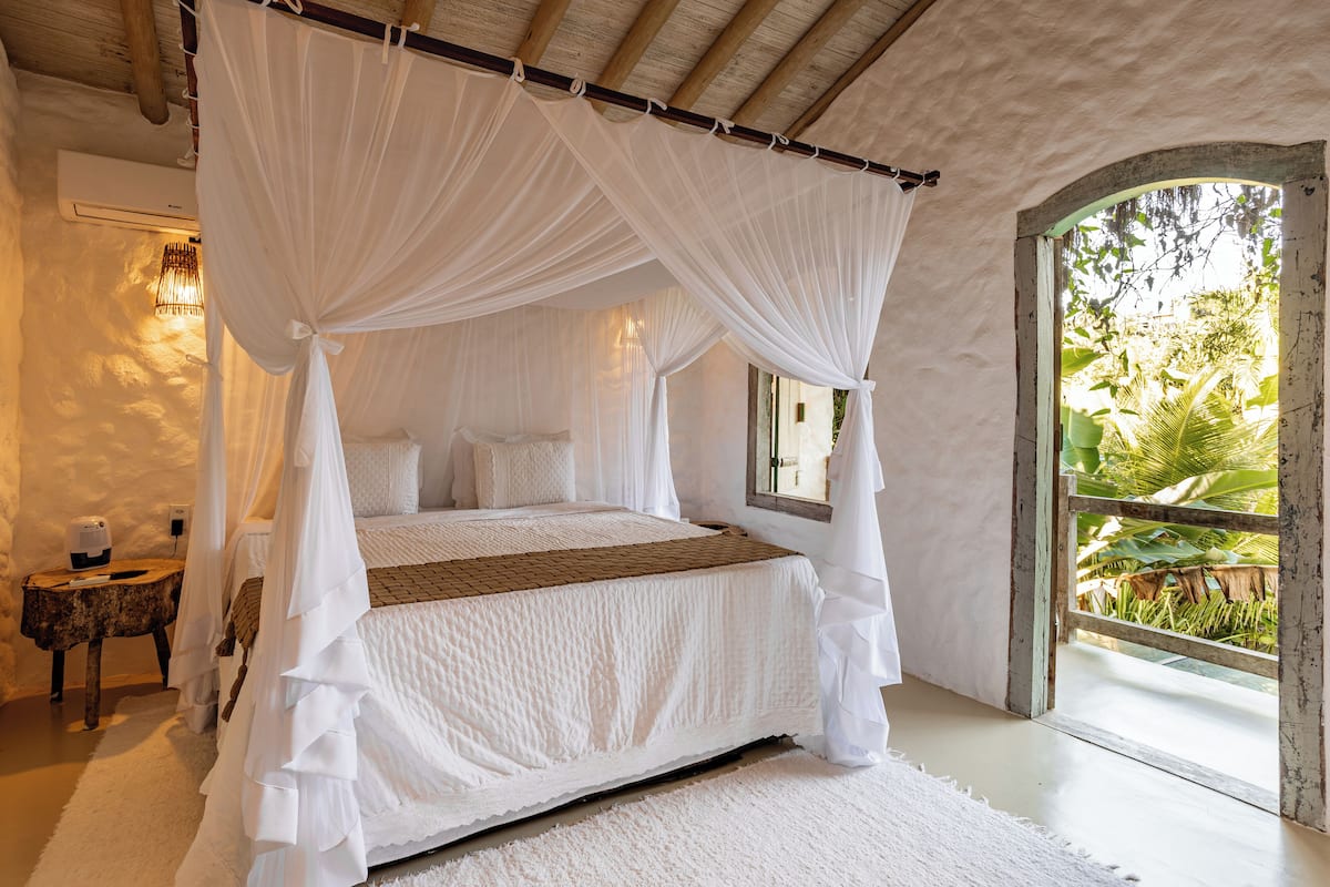 Un dormitorio con cama con dosel, una mesita de noche y vistas a la vegetación a través de una ventana.