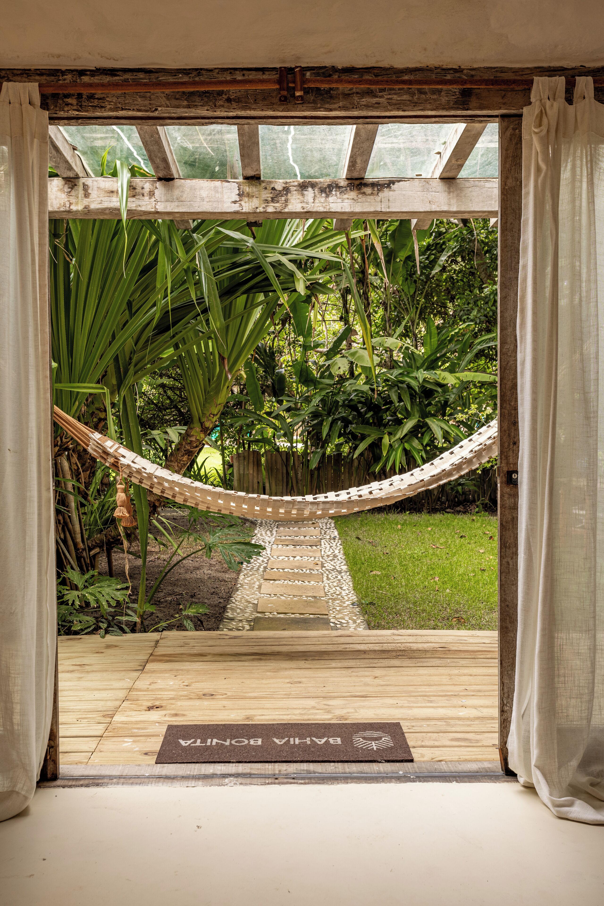 A wooden deck leading to a hammock, surrounded by lush greenery and a wooden pergola with a glass ceiling.