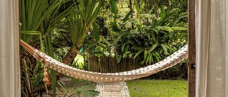 A wooden deck leading to a hammock, surrounded by lush greenery and a wooden pergola with a glass ceiling.