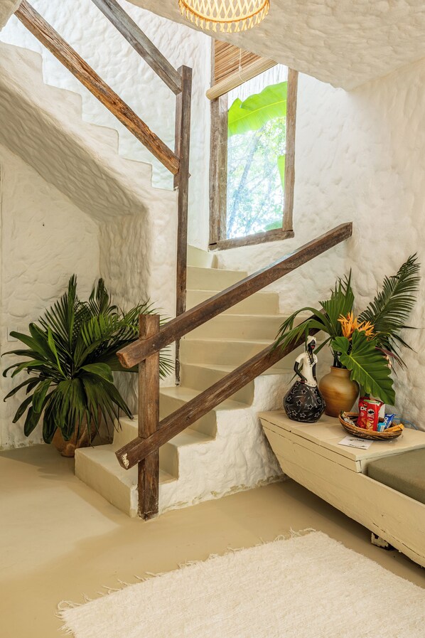 Una escalera con barandilla de madera, paredes blancas y un ventanal pequeño. Hay plantas en macetas y un objeto decorativo en el rellano.