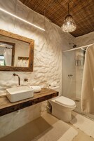 Un baño de diseño rústico con tocador de madera, un espejo grande, un lavamanos blanco, inodoro y ducha con mampara de vidrio.