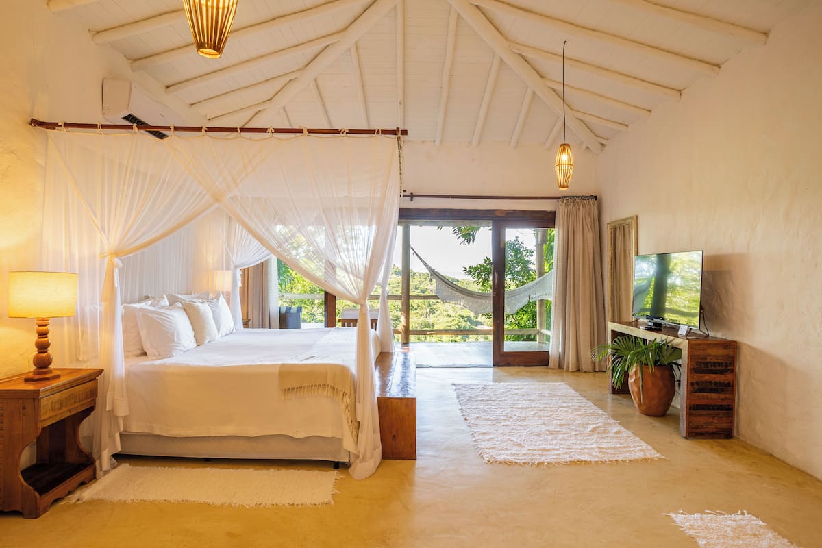 Un dormitorio con cama, televisión, mesita de noche, planta en maceta y vistas al exterior.