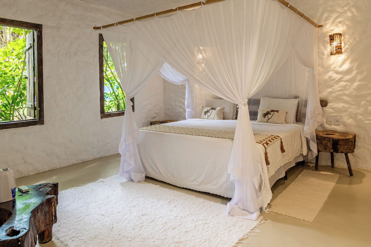 Un dormitorio con una cama con dosel, ropa de cama blanca, una alfombra blanca y una mesita de noche de madera.