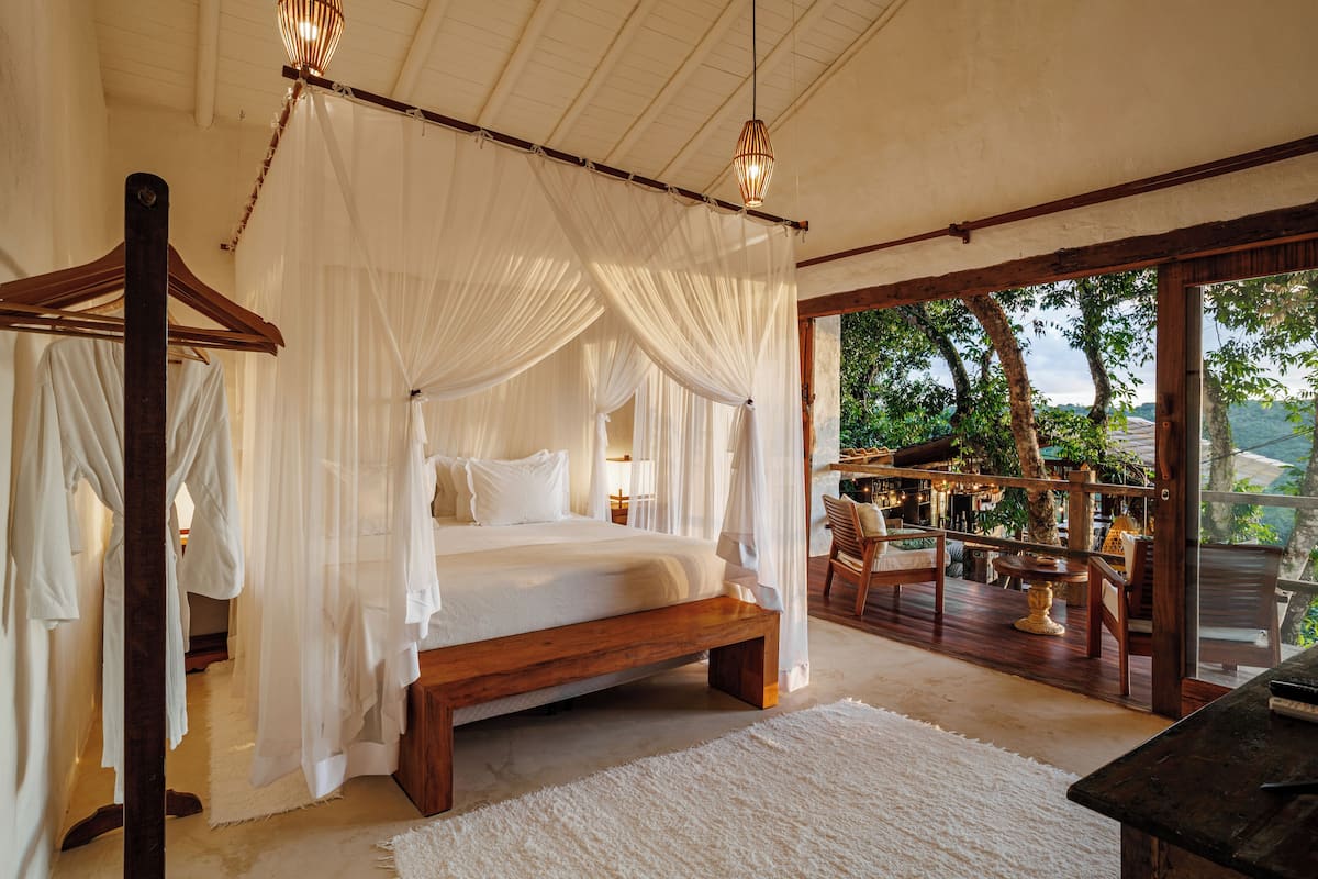 Un dormitorio amplio con una cama grande, cabecera de madera y dosel. Cuenta con un balcón con asientos exteriores y vistas del paisaje circundante.