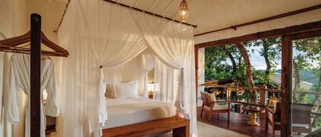 A spacious bedroom with a large bed, wooden headboard, and a canopy. There is a balcony with outdoor seating and a view of the surrounding landscape.