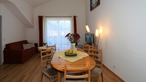 Un coin repas avec une table en bois, des chaises et un vase rempli de fleurs. On aperçoit un canapé rouge et une fenêtre avec des rideaux à l’arrière-plan.