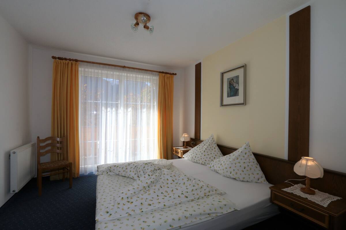A hotel room with a bed, a chair, a nightstand with a lamp, a window with curtains, and a framed picture on the wall.