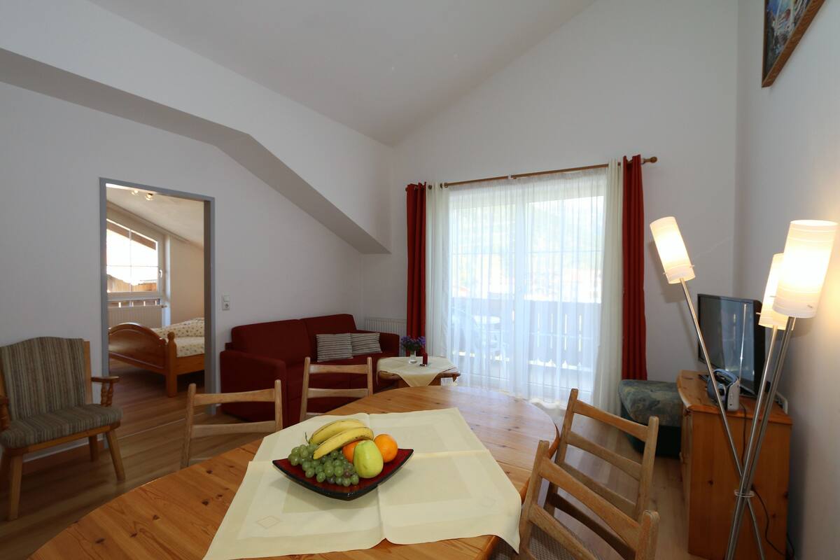 A room with a dining table set with fruits, a sofa, a TV, and a lamp.