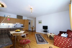 A living room with a sofa, a TV, a dining table with chairs, a kitchen area, and a hallway.