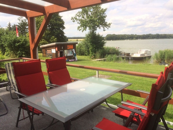 Ein Außenbereich mit Sitzgelegenheit, einem Tisch mit Glasplatte und roten Stühlen, mit Blick auf einen See mit Booten und einem kleinen Gebäude im Hintergrund.