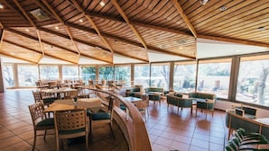 Een ruime binnenruimte met houten balken in het plafond, grote ramen en diverse zithoeken, waaronder tafels en stoelen.