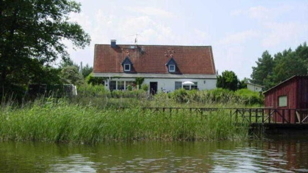 Una casa junto a un cuerpo de agua tranquilo, con techo rojo, rodeada de vegetación.
