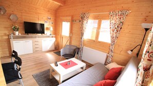 A cozy cabin with a living area featuring a sofa, a coffee table, and a TV. There is a kitchen with a white countertop and cabinets, and a dining area with a chair. - Cozy vacation home in beach location for 4 people and only 100m to the bathing area (Plau am See)