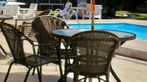 A poolside area with wicker chairs and white umbrellas, a pool, and a building in the background. - Pousada Belmare (Rio das Ostras)