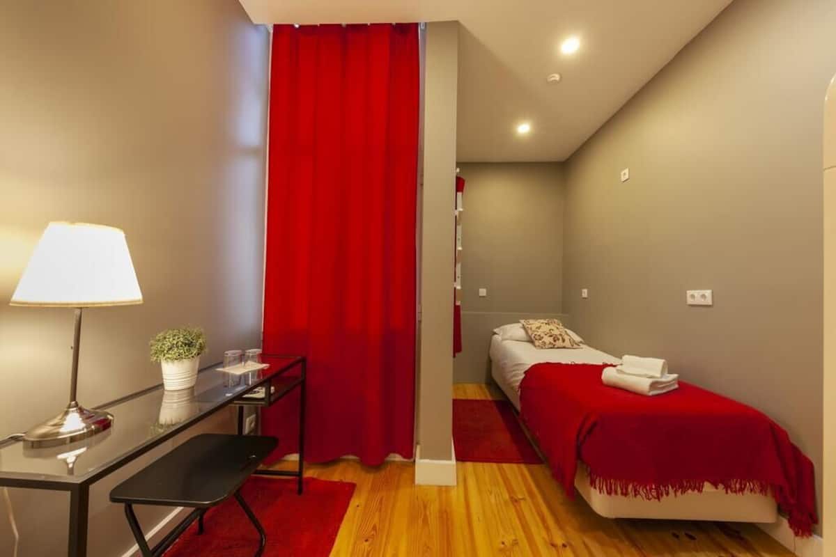A compact bedroom with a bed, a desk with a lamp, and a red curtain.