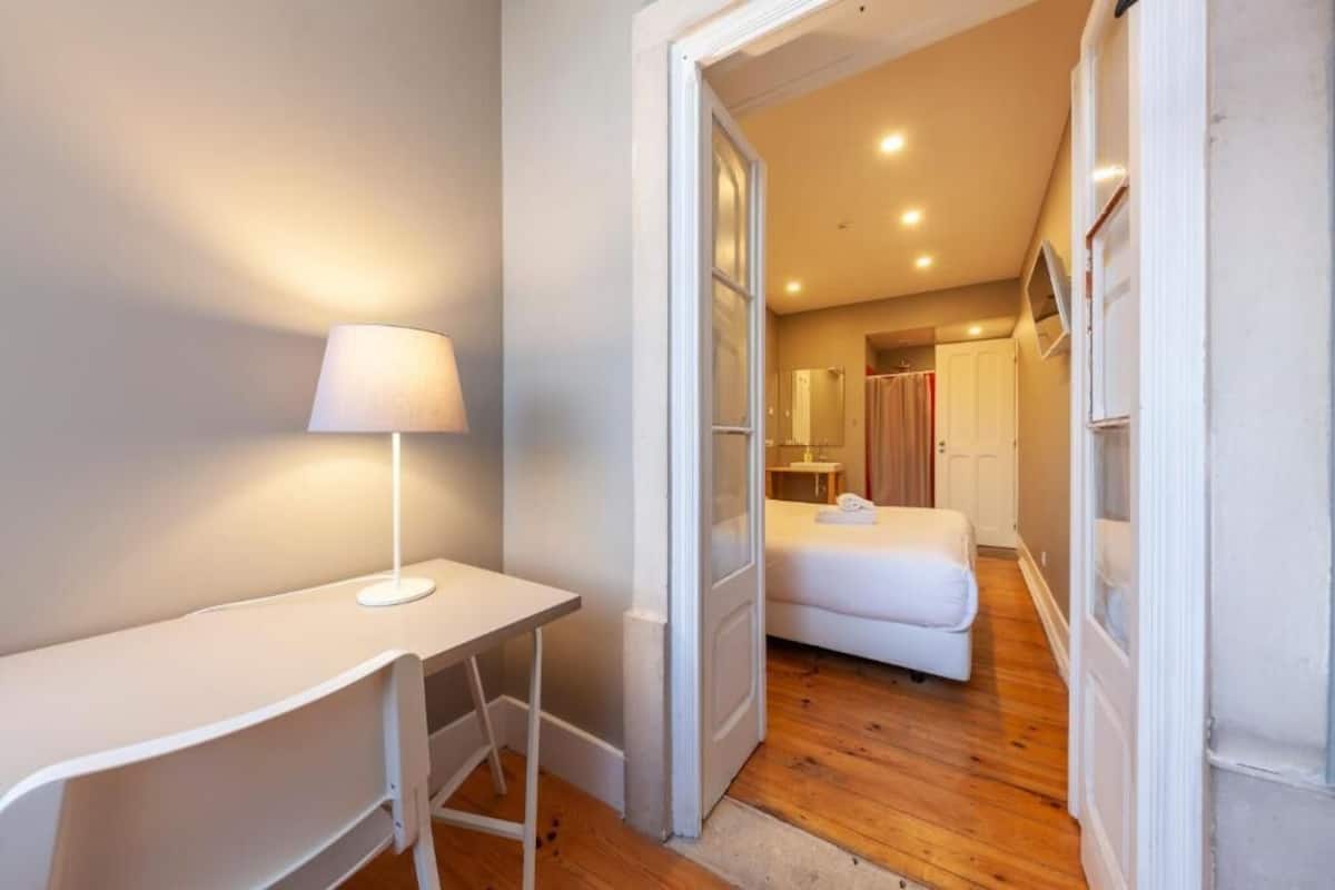 A modern, well-lit room with a white desk and lamp, a bed with a white duvet, and a wooden floor.